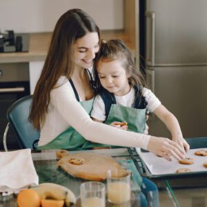 Mother, with daughter sitting on her lap, removes cookies from a cookie sheet pan in their kitchen