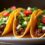 Three crispy beef tacos on a platter with Lettuce, Tomato and peppers